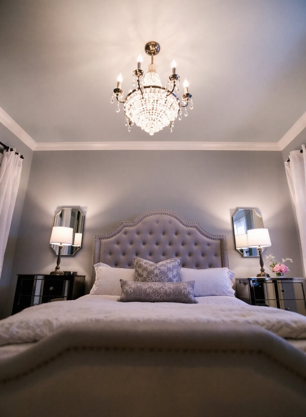 Cozy twilit 11x13ft bedroom with pale gray walls, light blue ceiling, upholstered silver-gray velvet bed, crystal chandelier, mirrored nightstands, and layered window treatments, shot from a low-angle.