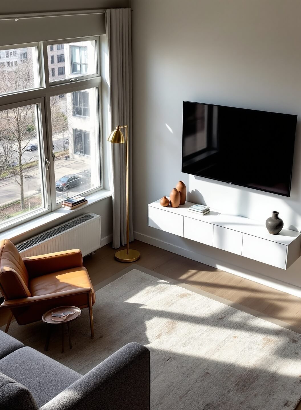 Modern, minimalist 12x18ft living room with floor-to-ceiling windows bathing the space in morning light, featuring a wall-mounted TV above a white media unit, a gray sofa with console table adorned with ceramics, a reading nook furnished with a cognac leather armchair and brass lamp, all captured from a high-angle view to emphasize spatial organization and clean lines.